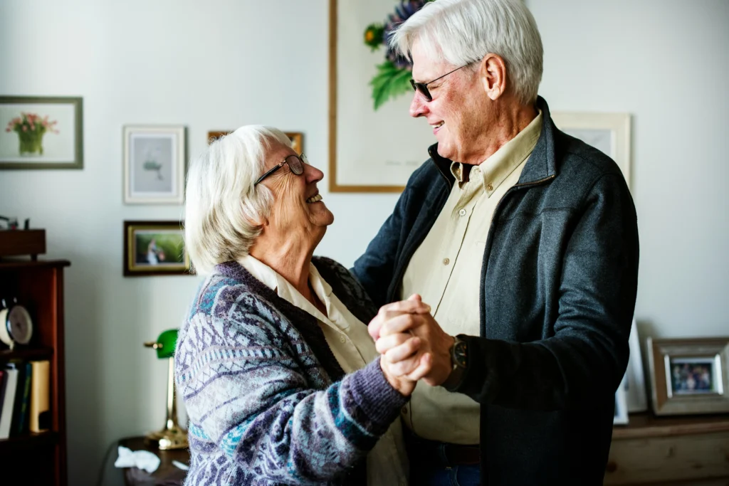Older couple smiling and holding hands while dancing together at home.